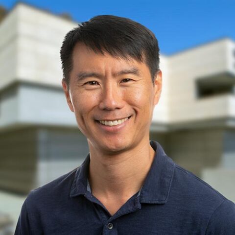 Professional headshot of Professor Andrew Su in front of a white building, wearing a blue t-shirt.