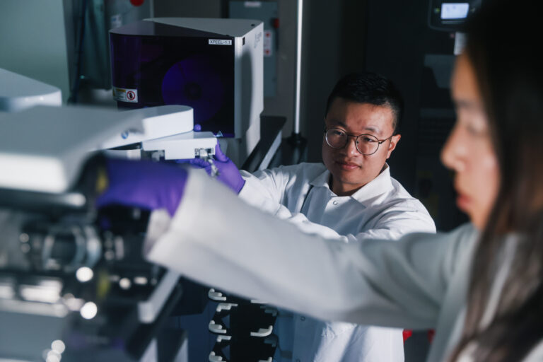 Calibr-Skaggs researcher in a white lab coat and purple gloves loading samples into a plate-reading instrument in a darkened laboratory.