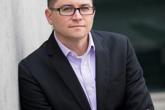 Professional headshot of Jared Machado, Scripps Research leadership, in a dark suit with glasses leaning against a concrete wall.