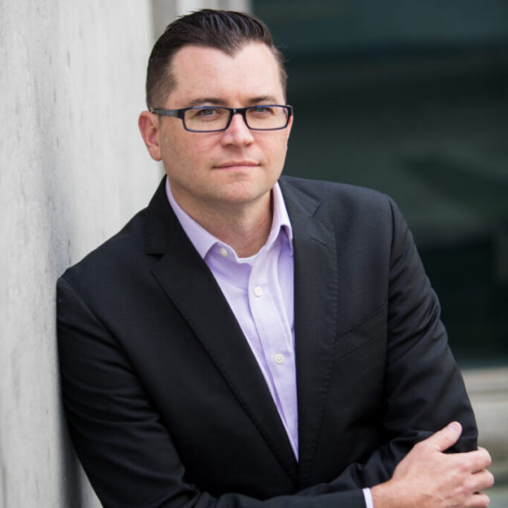 Professional headshot of Jared Machado, Scripps Research leadership, in a dark suit with glasses leaning against a concrete wall.