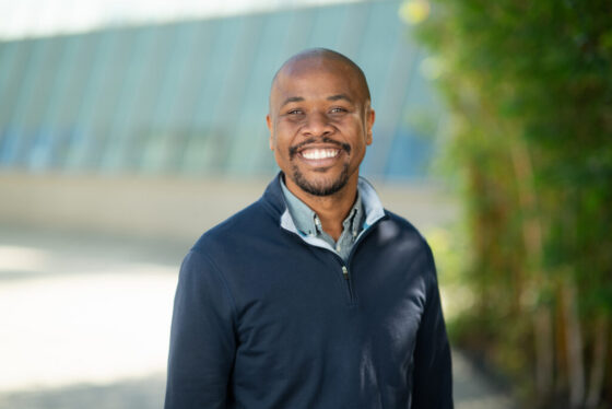 Professional headshot of Rohan Barrett, Scripps Research leadership, smiling broadly in a navy pullover outdoors with greenery in background.