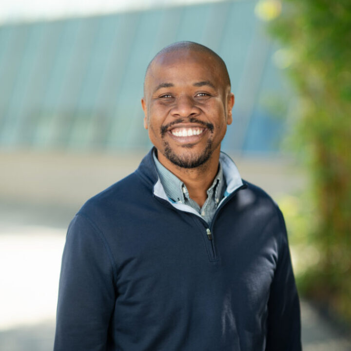 Professional headshot of Rohan Barrett, Scripps Research leadership, smiling broadly in a navy pullover outdoors with greenery in background.