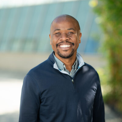 Professional headshot of Rohan Barrett, Scripps Research leadership, smiling broadly in a navy pullover outdoors with greenery in background.