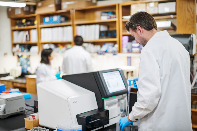 Scripps Research scientists in white lab coats operating a DNA sequencing instrument in a human genomics laboratory.