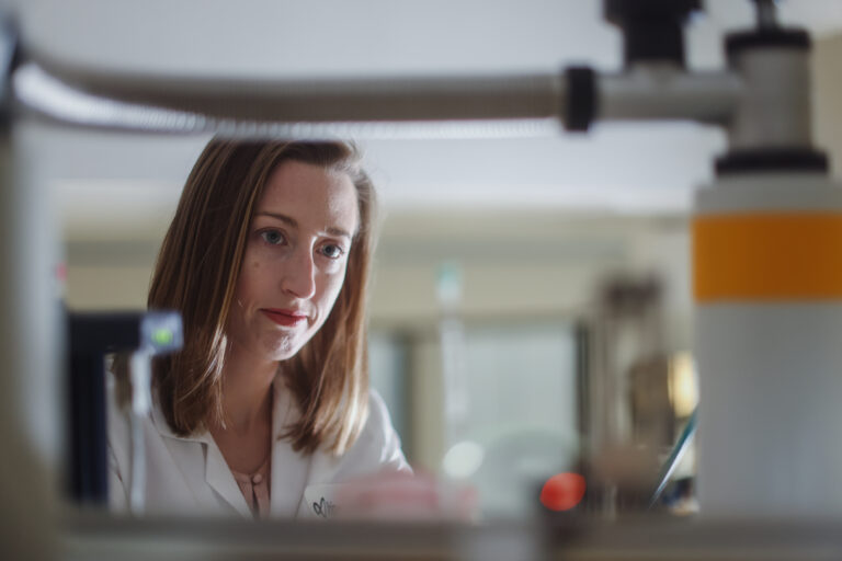 Scripps Research Fellow in a white lab coat examining samples at a laboratory bench framed through scientific equipment.