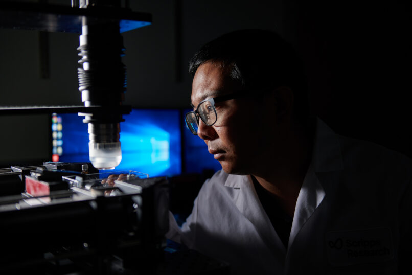 Scripps Research Fellow in a white lab coat and glasses working at a fluorescence microscope in a darkened laboratory.
