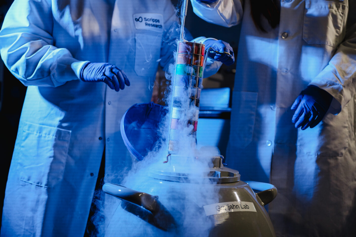 Two Scripps Research scientists in lab coats and blue gloves removing a sample rack from a liquid nitrogen cryogenic storage dewar in a blue-lit laboratory.