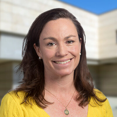 Professional headshot of Katja Lamia, Scripps Research faculty member, in a yellow cardigan with a campus building background.
