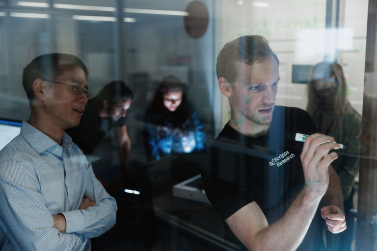Man writing on a glass wall with another man observing.