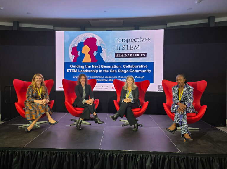 Four women sitting in red armchairs on a stage.