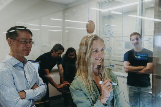 Student writing on glass wall with colleagues observing.